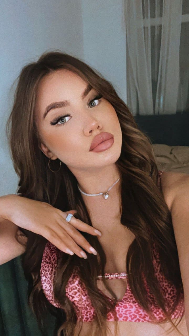Woman in a pink leopard bralette and jewelry poses indoors near sheer curtains and a bed.