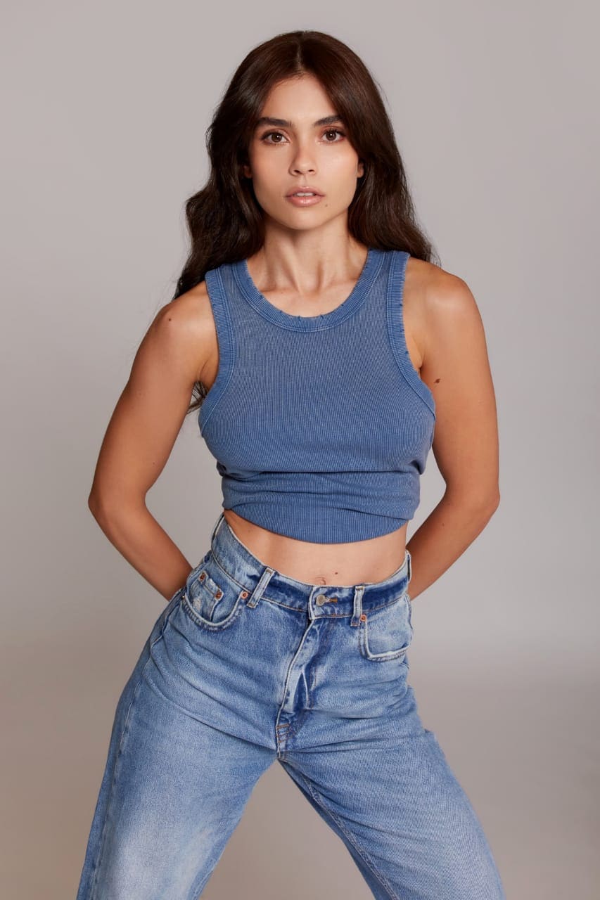 Emy stands with hands on hips in a blue ribbed crop top and light-wash high-waisted jeans against a gray studio background.