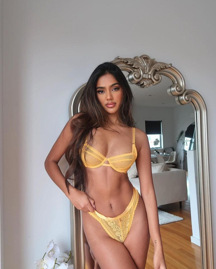 Ilse stands before an ornate mirror in a yellow lace two-piece set, hand on hip, against a white wall and wooden floor.