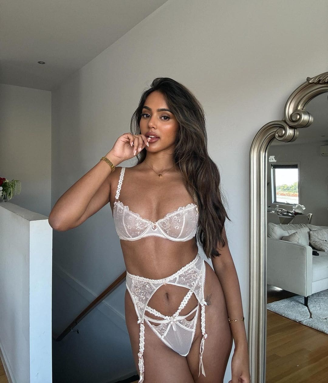 She wears a white lace bra and high-cut briefs with side ties, hand raised to mouth, beside a large mirror in a bright room.