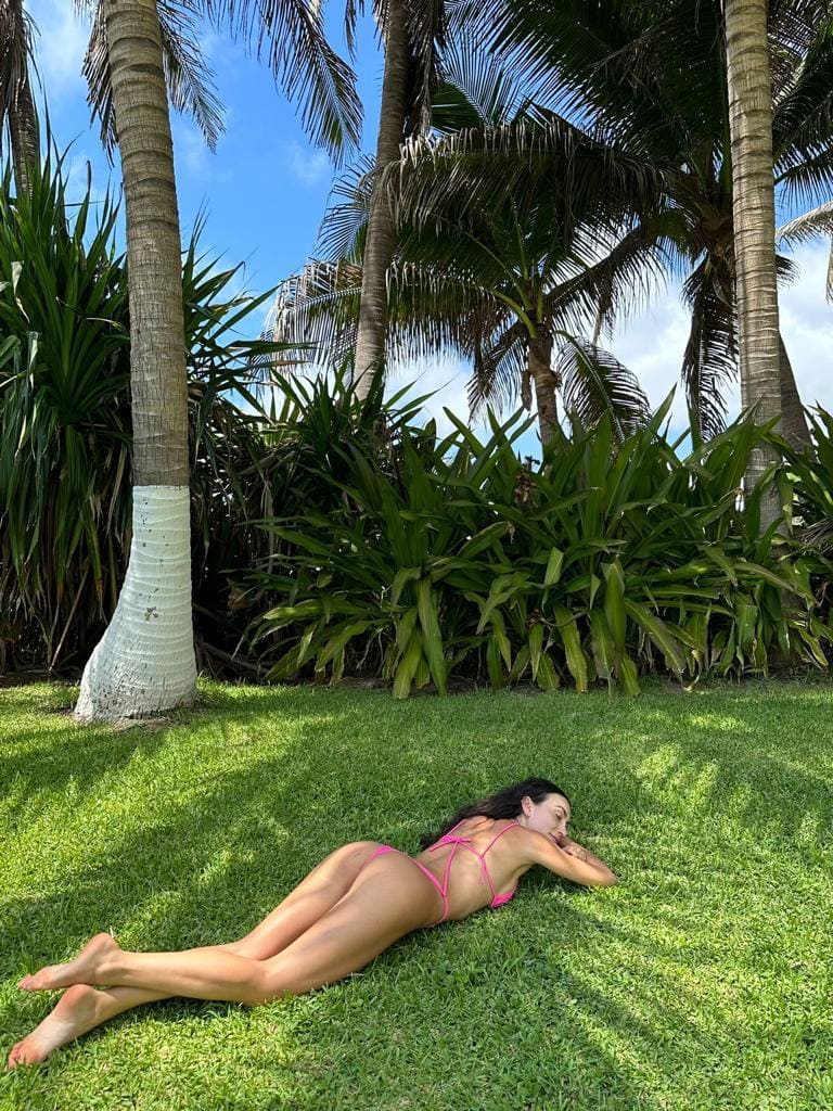 A woman lies on her stomach on green grass in a pink string bikini, arms under head, palms behind.