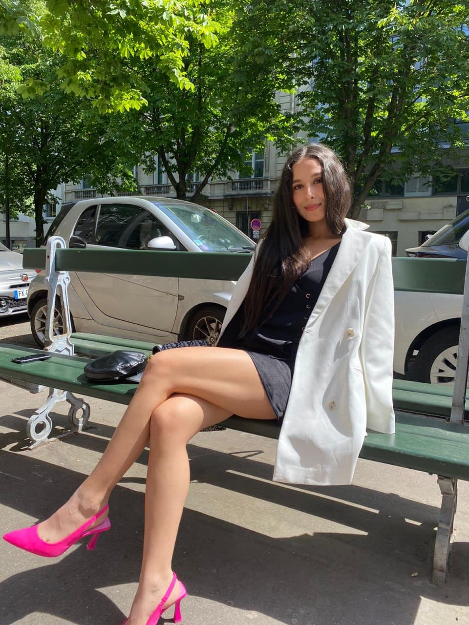 Salome sits on a green park bench in a white blazer, black top, short skirt, and pink heels, with cars and trees behind her.