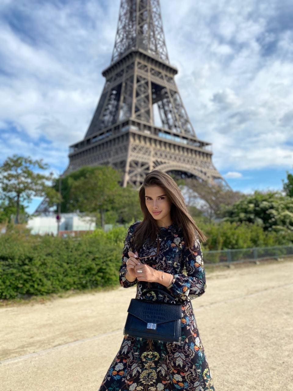 Standing outdoors before an iron lattice tower, she wears a floral dress and holds a dark handbag at her waist.