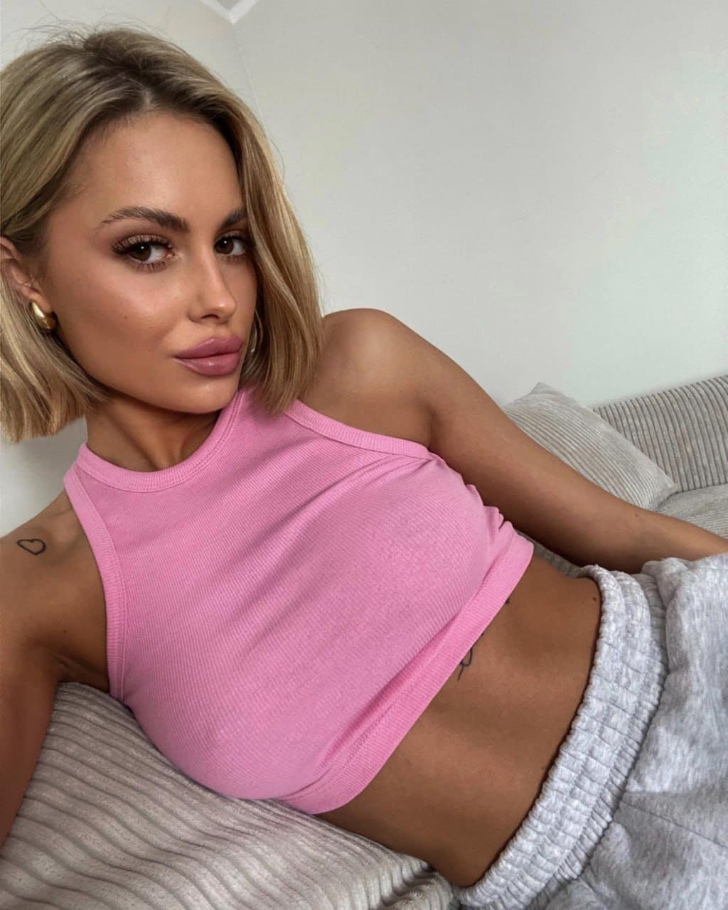 A blonde woman in a pink crop top and grey sweatpants lies on a couch, showing a heart tattoo on her shoulder.