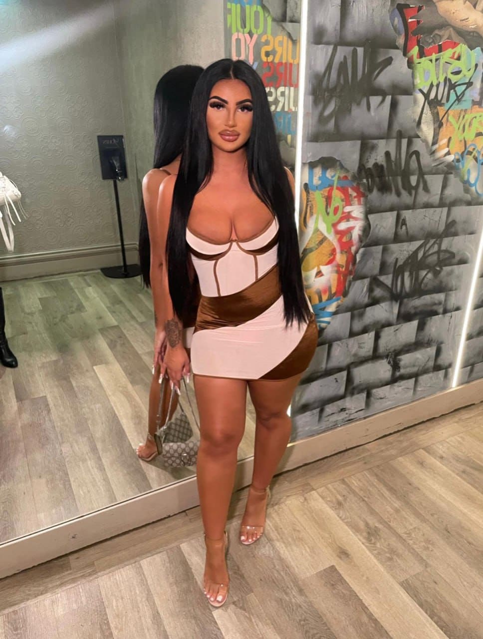 Shannon stands before a mirror in a short dress with cut-outs and heels, set against a graffiti wall and wooden floor.