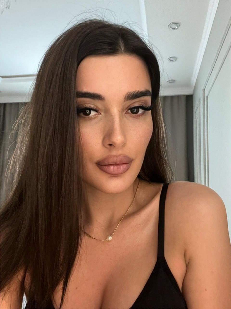 Amina poses in a close-up selfie wearing a black spaghetti-strap top and gold necklace against white walls with recessed lights.