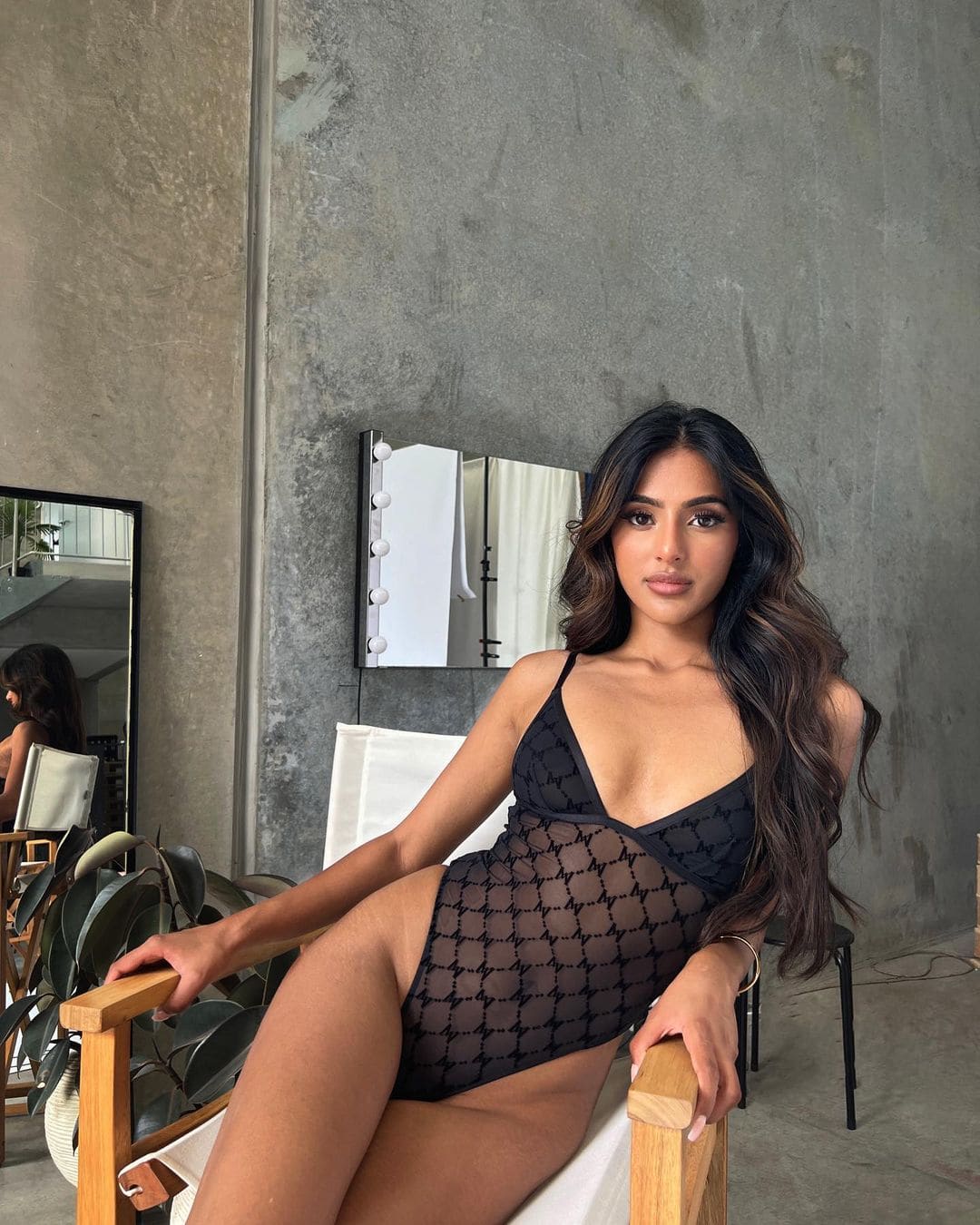 Sitting in a wooden chair against a concrete wall, she wears a black sheer patterned bodysuit with a high leg cut.