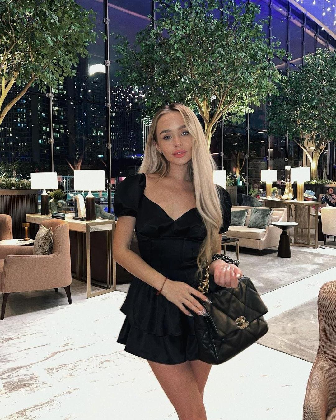 A woman in a short black dress with puffed sleeves and a sweetheart neckline stands in a modern lounge at night, holding a quilted black handbag.