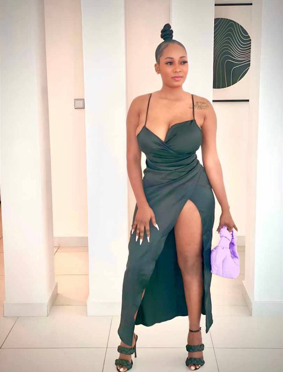 Camilla stands indoors in a dark green satin slip dress with a thigh slit, holding a light purple handbag and wearing green strappy heels.