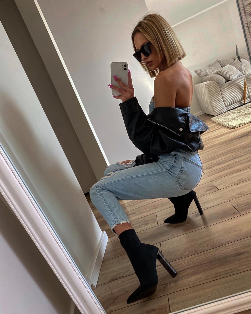 A woman in sunglasses, an off-shoulder leather jacket, and ripped jeans takes a mirror selfie on a wooden floor.