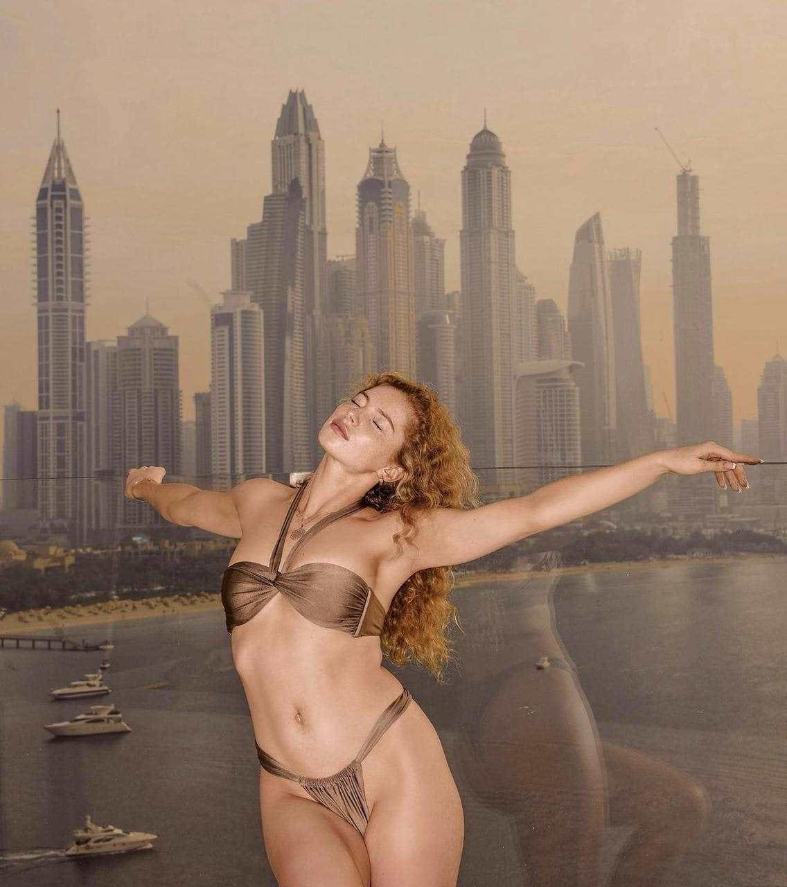 A woman with long curly hair stands in a brown bikini with arms outstretched before a painted city skyline backdrop.