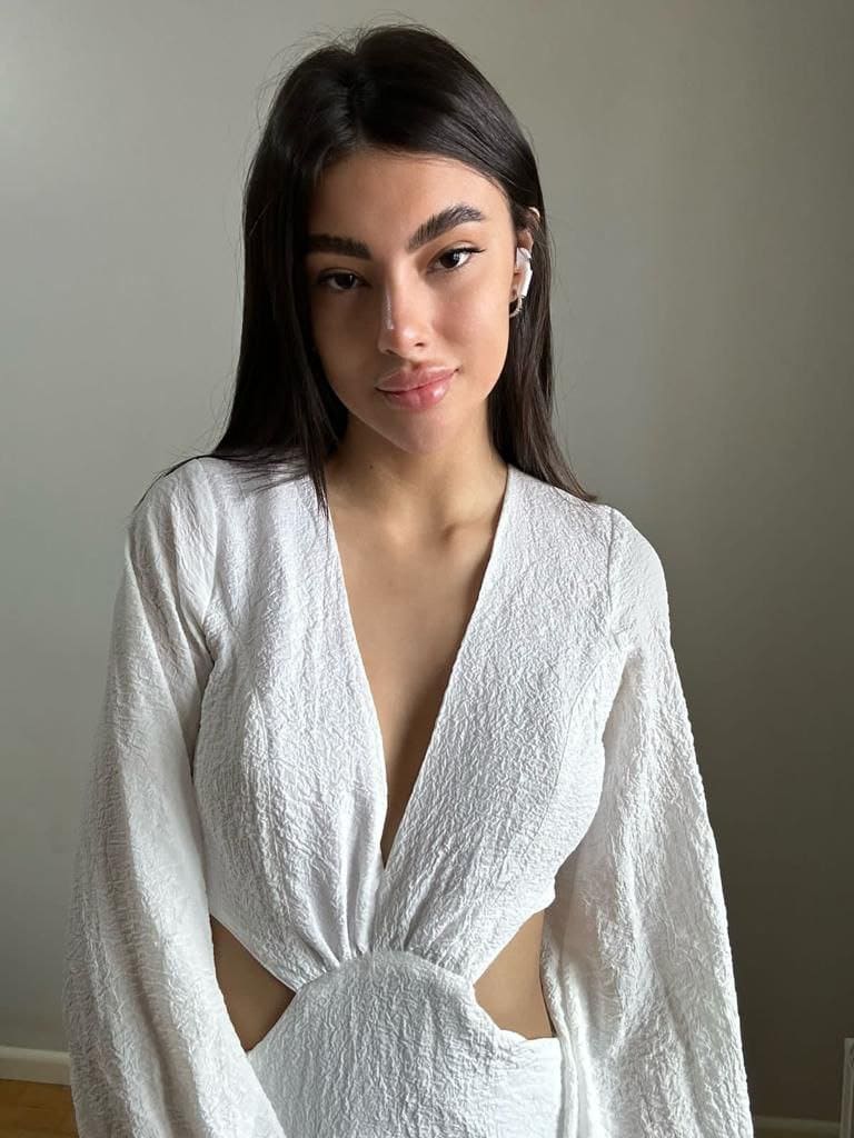 A woman in a white textured wrap top with side cutouts wears a wireless earbud, standing against a light wall.