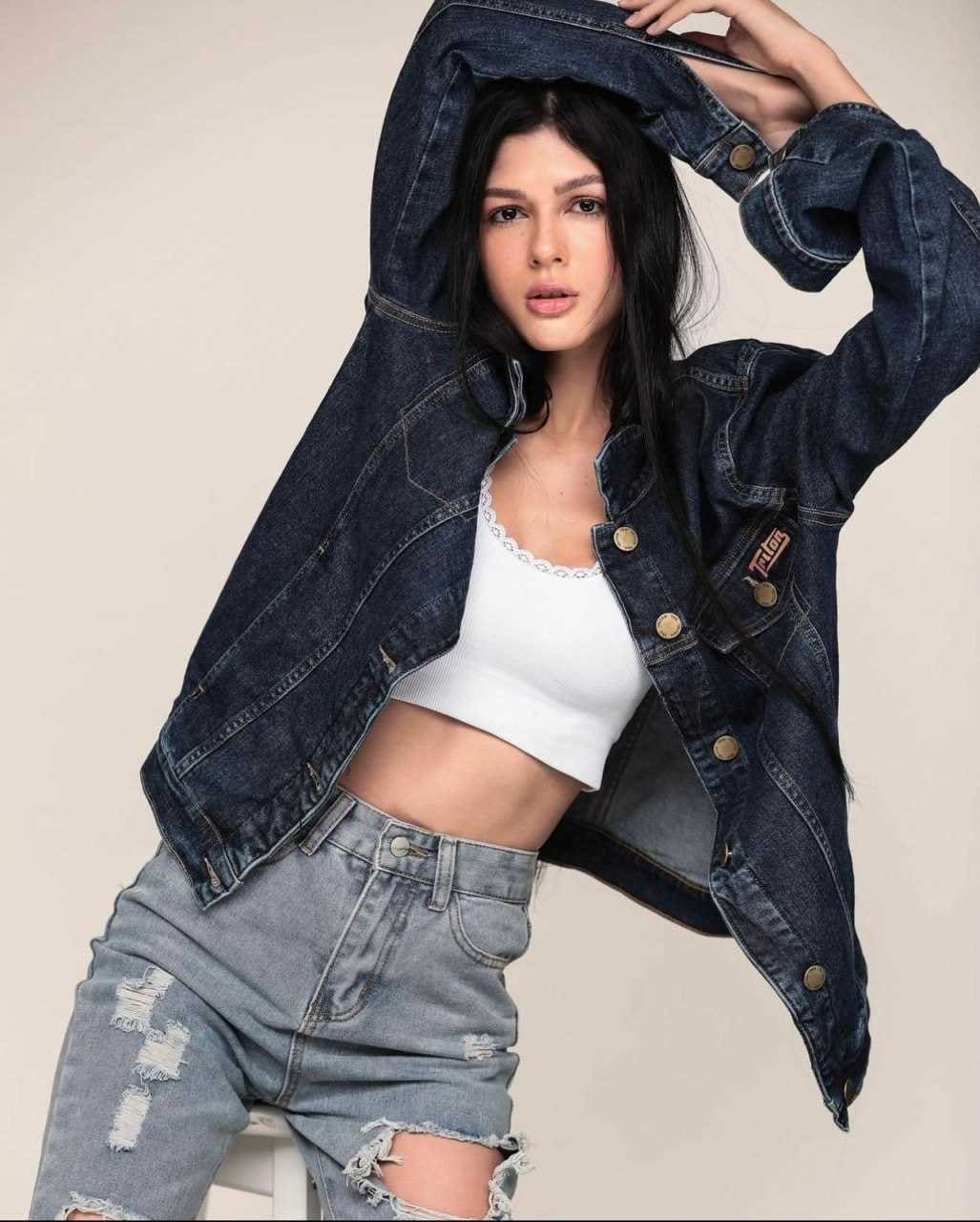 A woman in a white crop top and denim shorts poses in a studio, holding a dark denim jacket over her head with one arm raised.