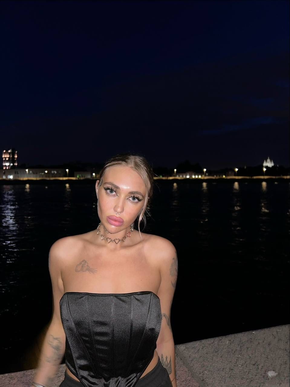 At night by water, a woman with hair pulled back wears a black strapless satin top and heart choker, showing chest and arm tattoos.