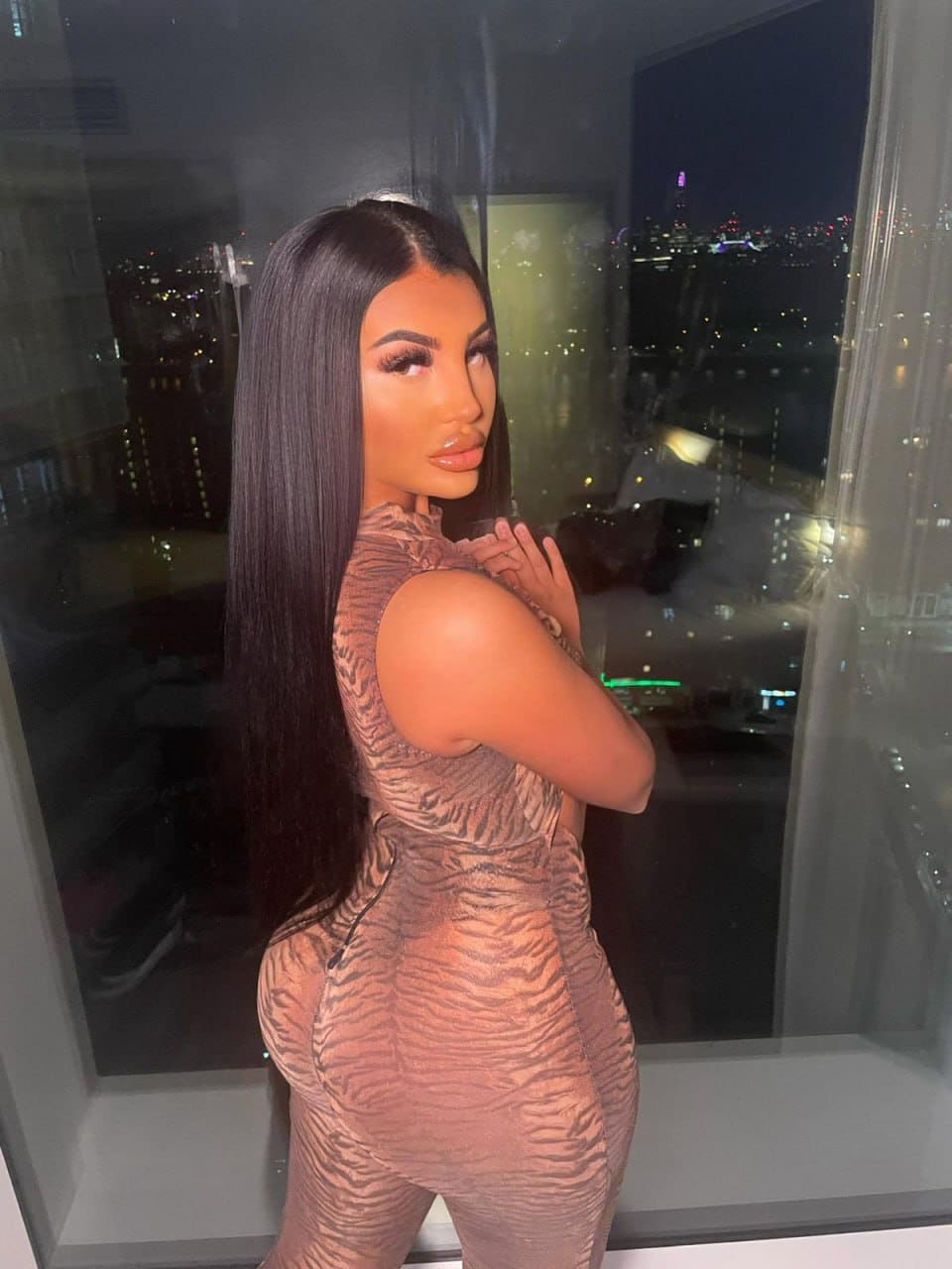 Profile view of a woman in a brown and black animal print dress, hands clasped, standing by a window overlooking a city at night.