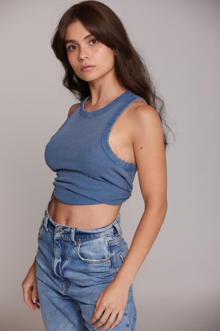 A woman in a three-quarter pose wears a blue ribbed crop top with shoulder cut-outs and light-wash jeans against a gray backdrop.