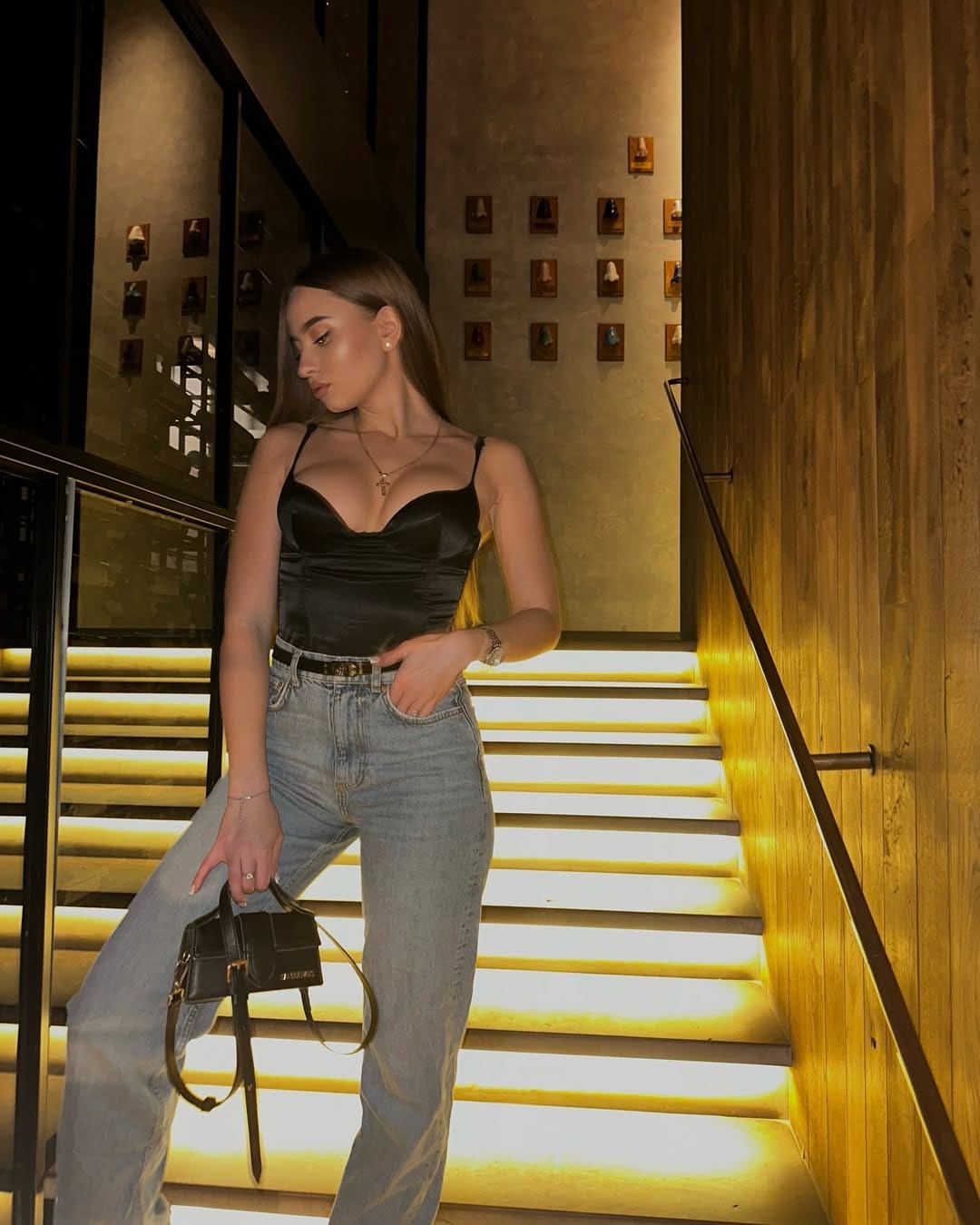 A woman stands on lit stairs in a black satin camisole and jeans, holding a handbag with one hand in her pocket, against a wall of framed items.