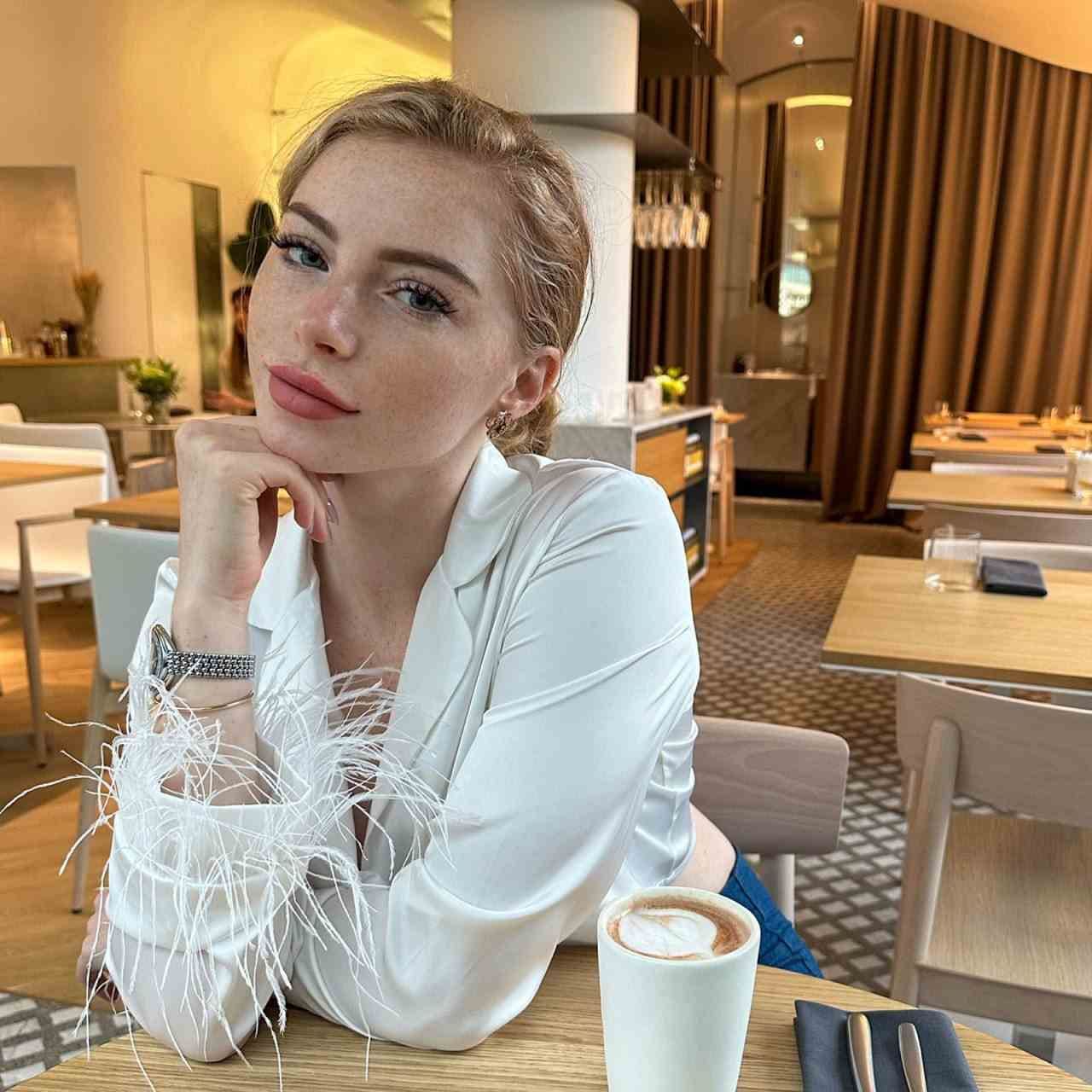 Sonya sits at a restaurant table in a white blouse with feathered cuffs, resting her chin on her hand beside a coffee cup.