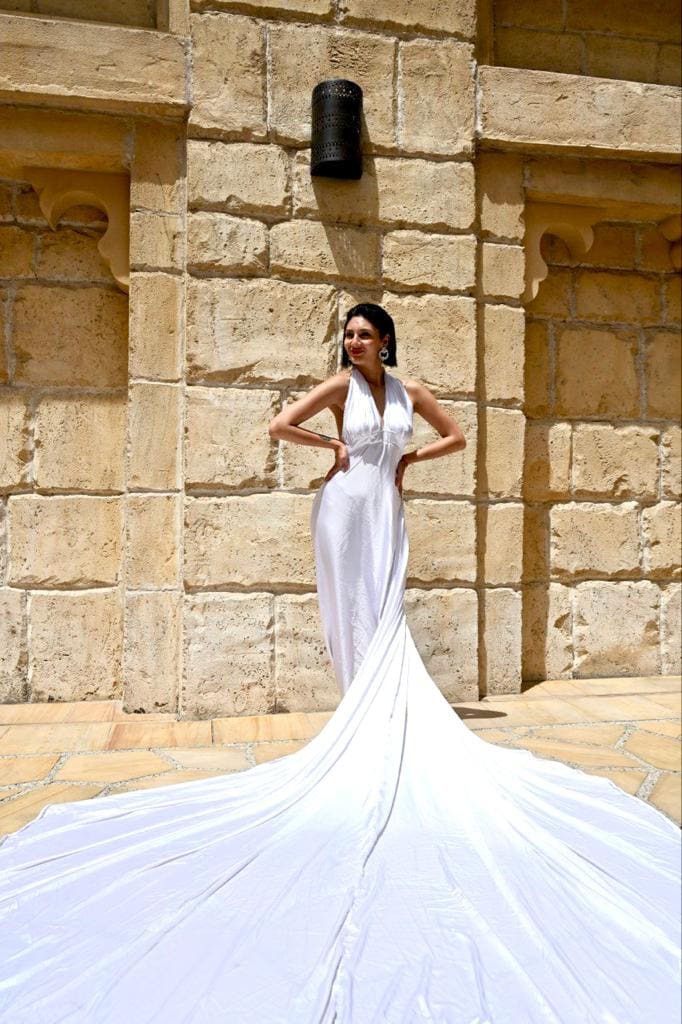 A woman in a long white halter gown with a train stands hands on hips before a tan stone wall, posing on paved ground.