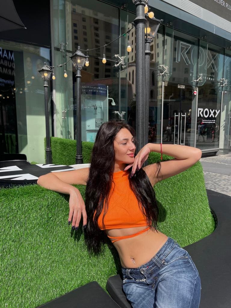A woman in an orange cropped tank and blue jeans sits on a black bench by a green planter, one arm resting on the edge.