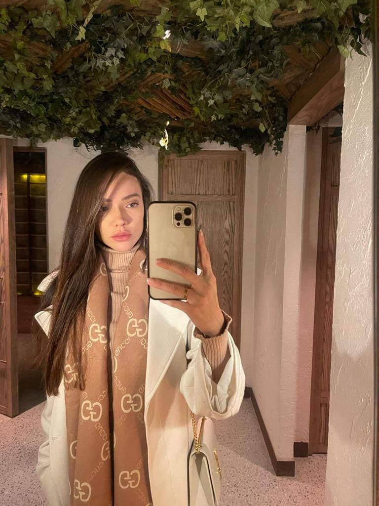 A woman in a beige turtleneck, white coat, and patterned scarf takes a mirror selfie in a hallway with wooden doors.
