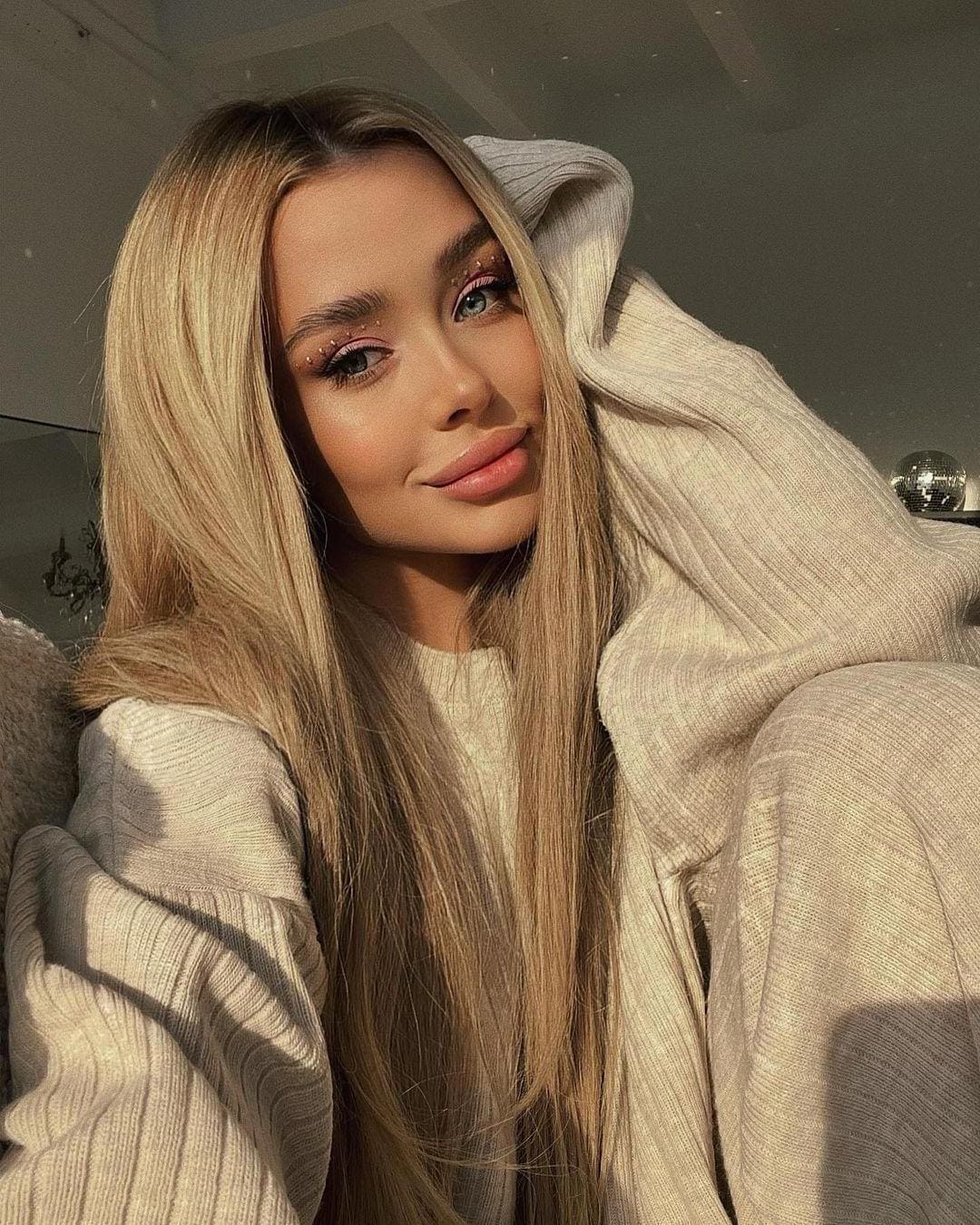 A woman with long straight blonde hair wears a cream ribbed knit sweater with the hood pulled up over one side of her head, lit by warm indoor light.