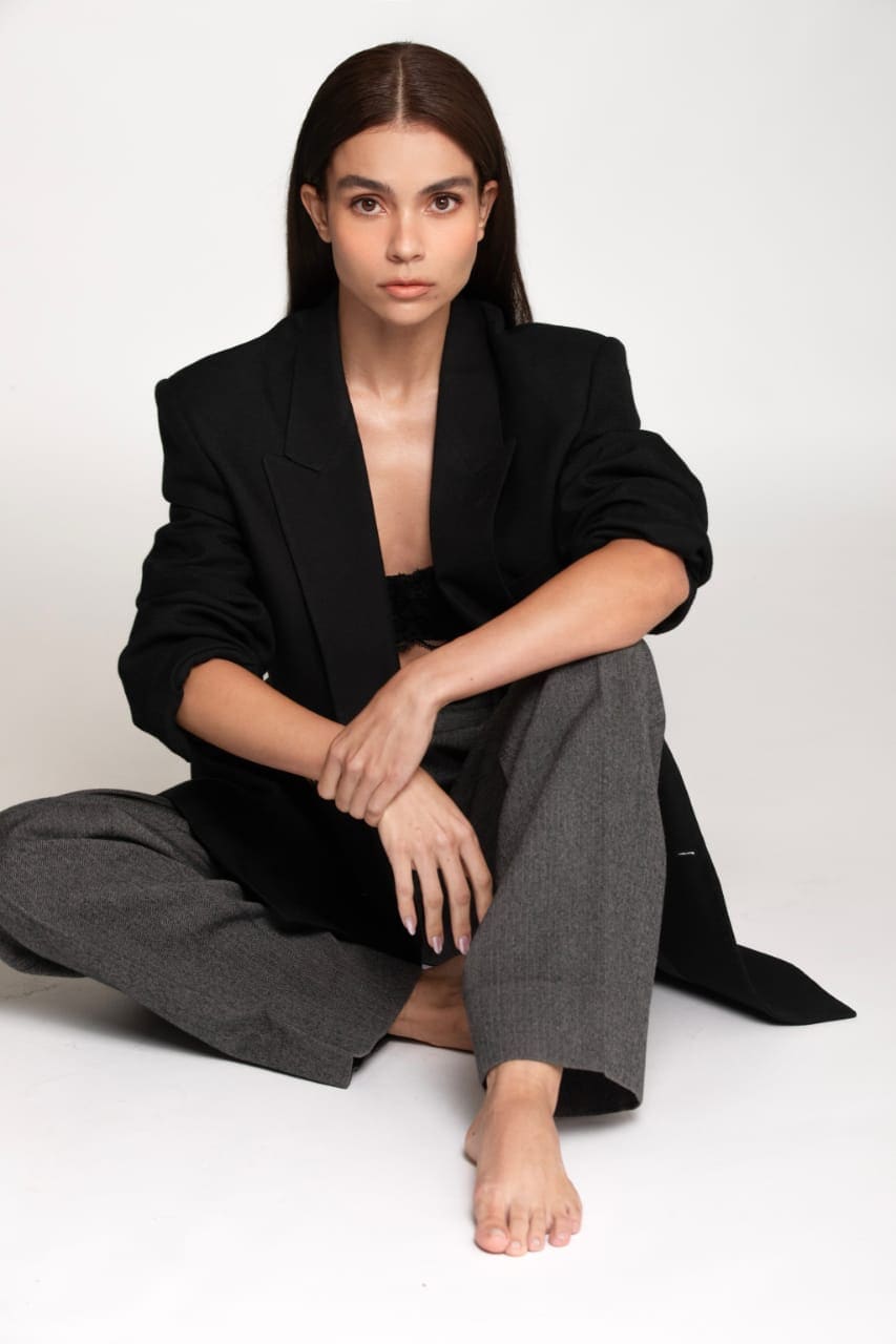 A woman sits cross-legged on a white floor in an open black blazer, black top, and grey trousers, with bare feet visible in front.