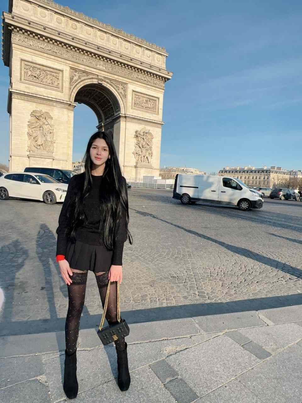 A woman in a black outfit and sheer tights stands on a cobblestone plaza holding a black handbag, with a large stone arch monument behind.