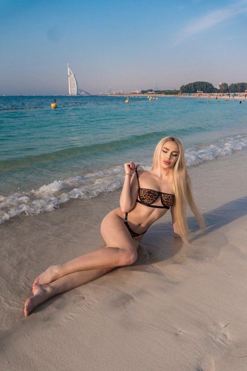 A blonde woman sits on wet sand in a leopard-print bikini, holding a strap, with the ocean and a distant sail-shaped building behind her.