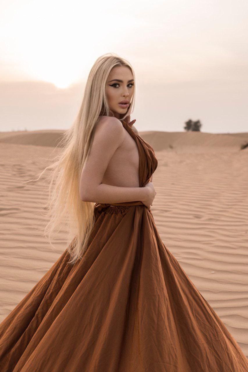 A blonde woman stands in a desert wearing a floor-length rust-colored gown with an open back, arms crossed and looking over her shoulder.