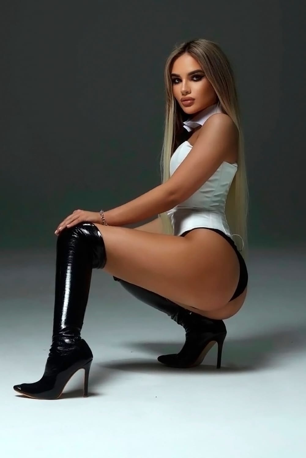 A blonde woman crouches in a white collared bodysuit, black briefs, and stiletto thigh-high boots, hand on her knee.