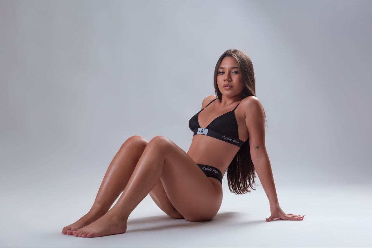 Sitting on a gray floor in a black bikini with logo bands, her legs are bent and one hand rests on the ground behind her.
