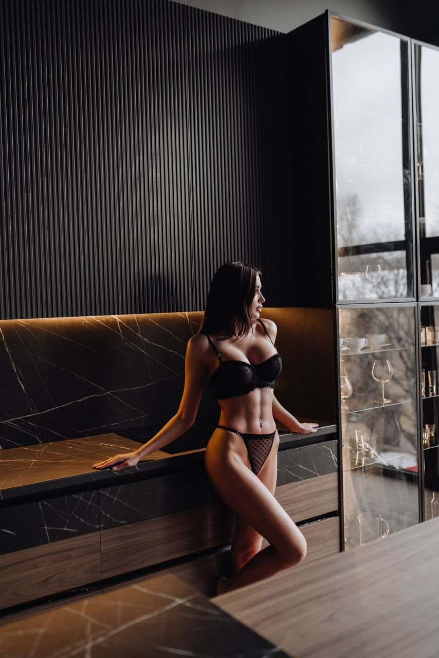 Leaning on a marble counter in a modern interior, she wears a black lace bra and sheer mesh briefs beside a glass cabinet.