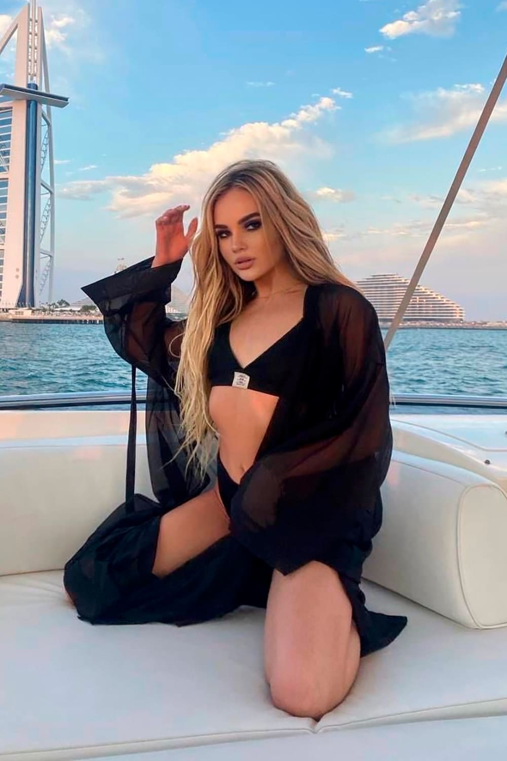 A blonde woman kneels on a boat in a black bikini and sheer robe, one hand raised, with water and a city skyline behind her.