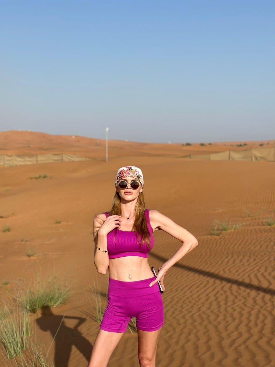 A blonde woman in a magenta sports bra, shorts, and headscarf poses in a desert landscape.