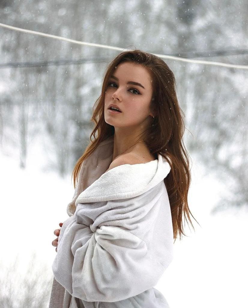 Maria stands in snow wrapped in a white blanket with one shoulder bare, blurred trees behind.