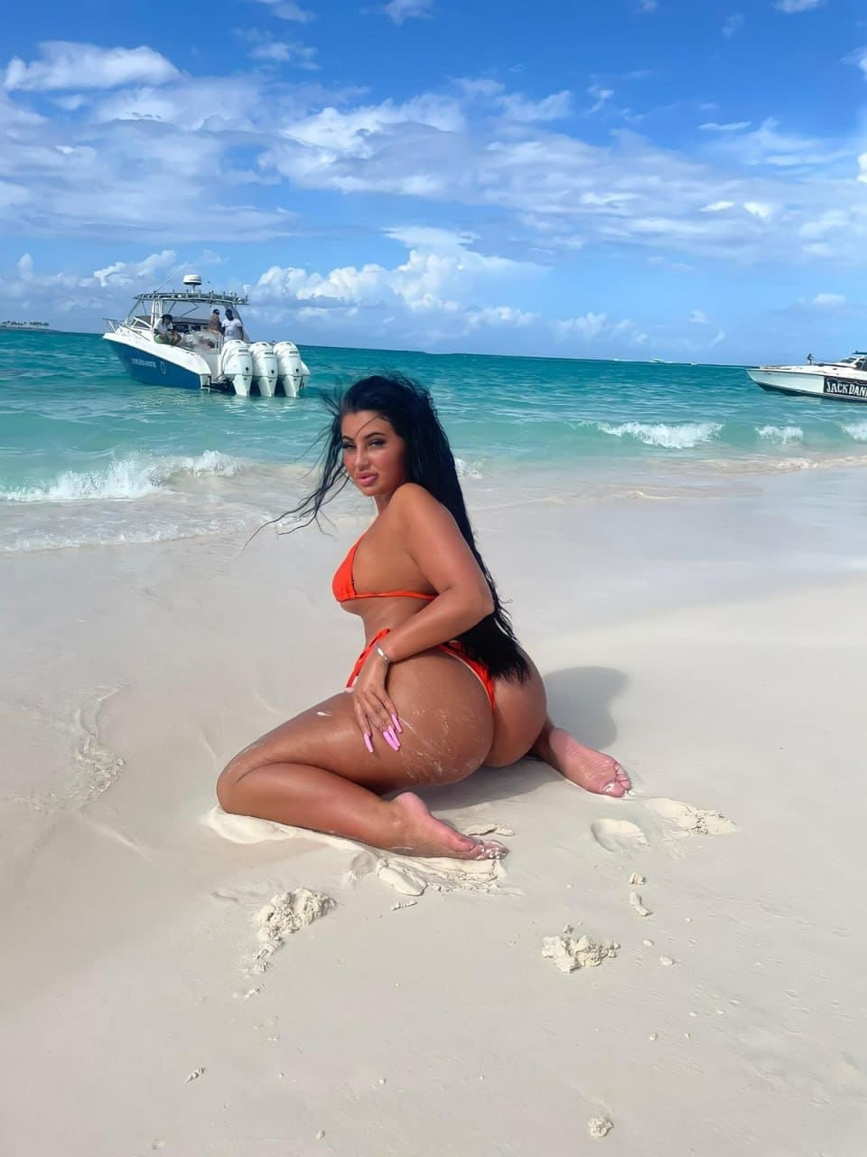 A woman in a red bikini kneels on a sandy beach with turquoise water and two boats visible under a partly cloudy sky.