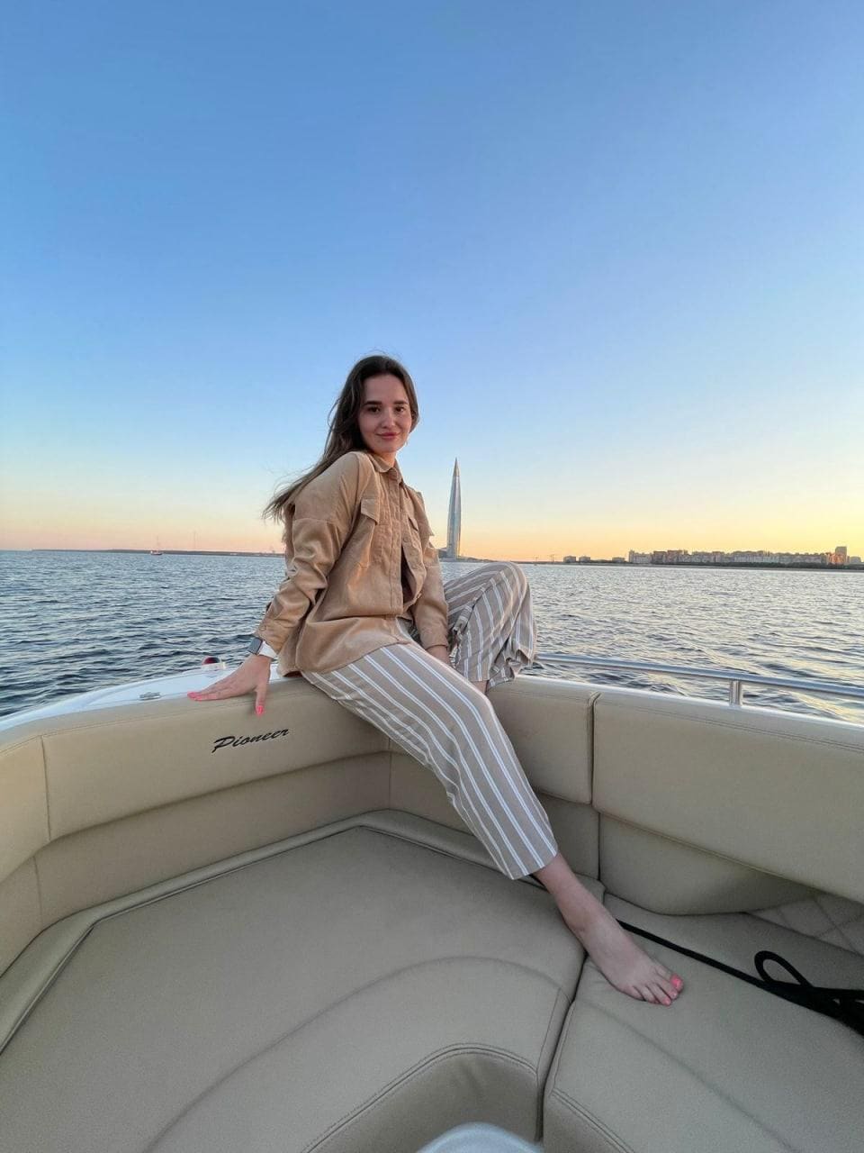 A woman sits barefoot on boat cushions wearing a tan shirt and striped trousers, with water and a distant skyline behind her.