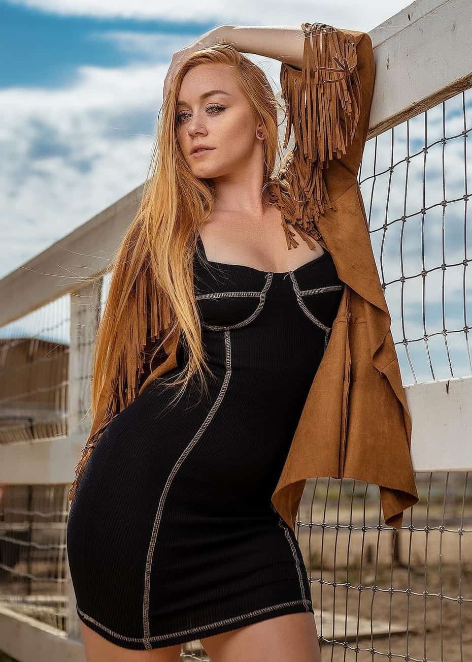 Blonde woman leans against a wire fence outdoors in a black mini dress and fringed suede jacket.