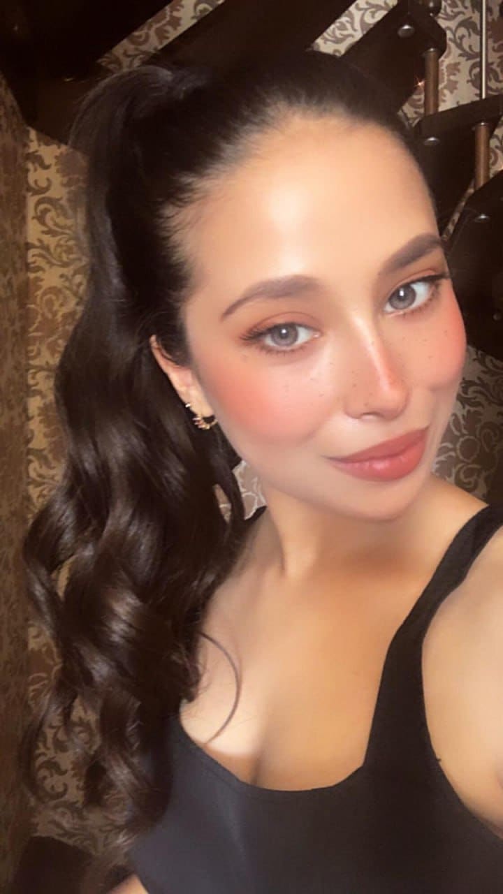 A close-up selfie of a woman with a high ponytail and black sleeveless top, posing against patterned wallpaper.