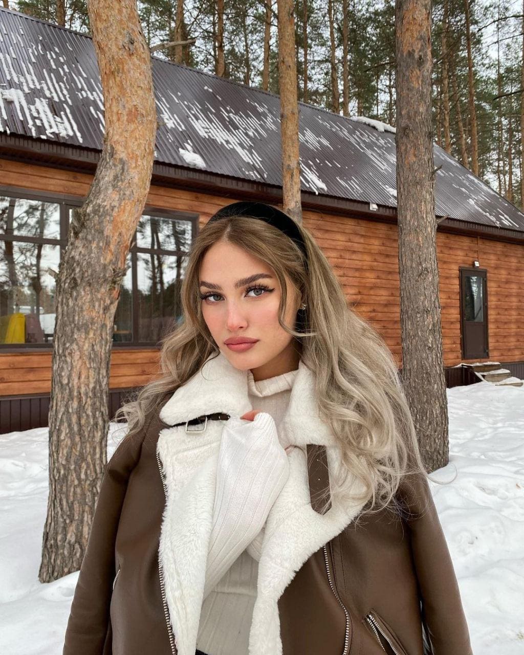 A woman in a brown shearling-collar jacket and cream turtleneck stands in the snow before a wooden cabin.
