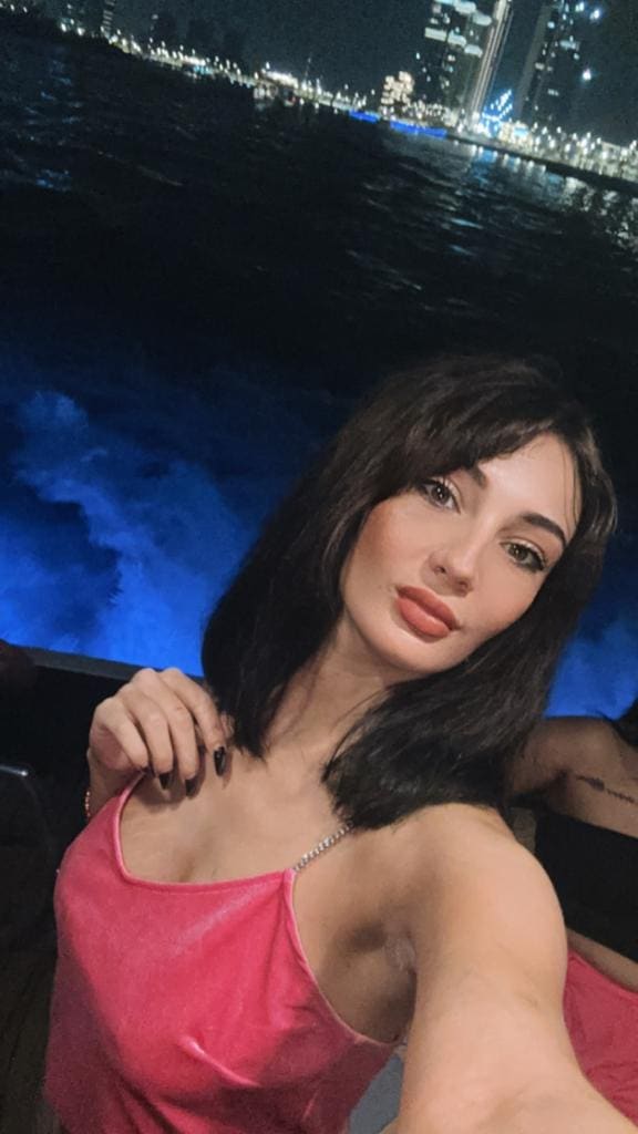 A selfie of a woman in a pink top on a boat at night, with a lit city skyline visible across the water behind her.