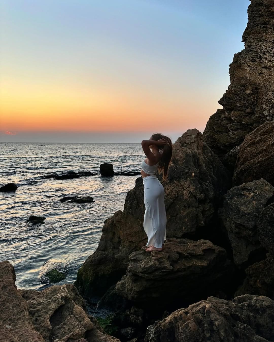 A barefoot woman in a white crop top and long skirt stands on a rocky sea outcrop at sunset, hands raised to her head.