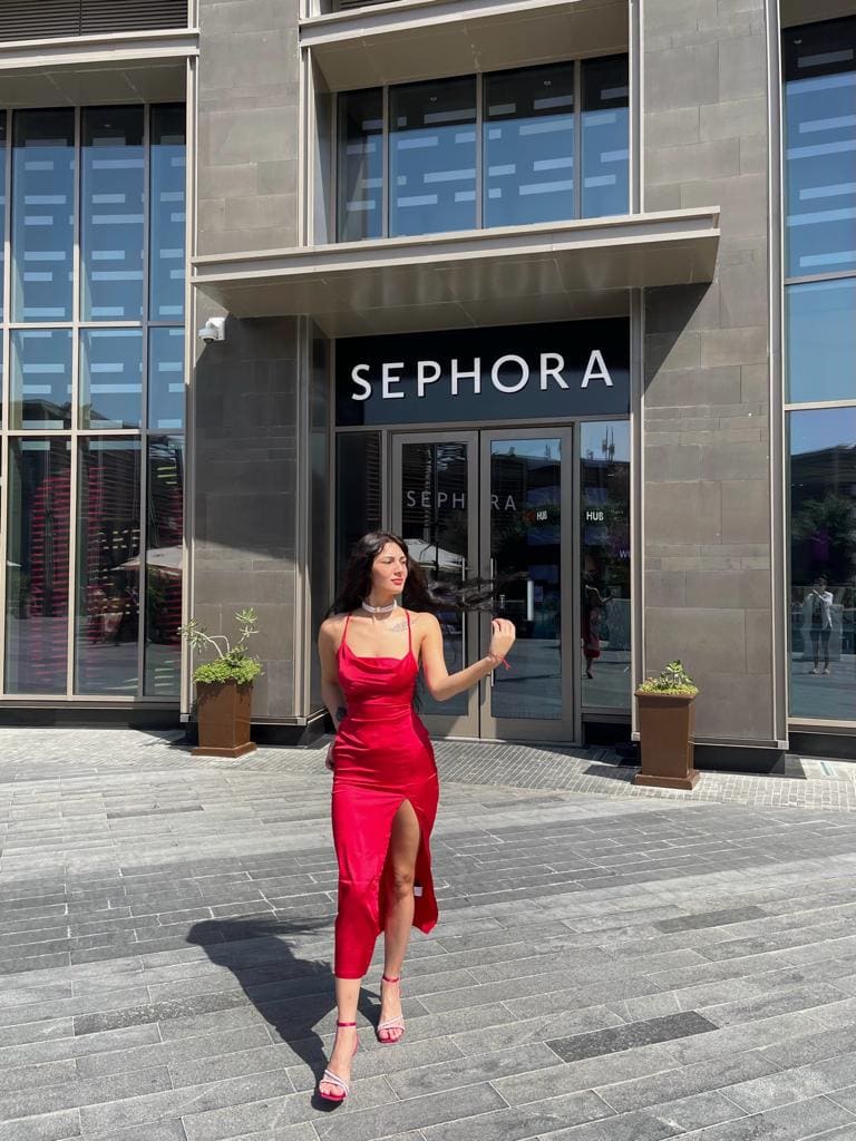 A woman in a red satin slip dress with a thigh slit stands on a paved plaza before a storefront, hand raised near her shoulder.