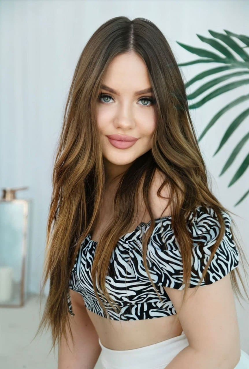 Milana wears a zebra-print off-the-shoulder crop top and white bottoms, posing by a white wall with a palm leaf.