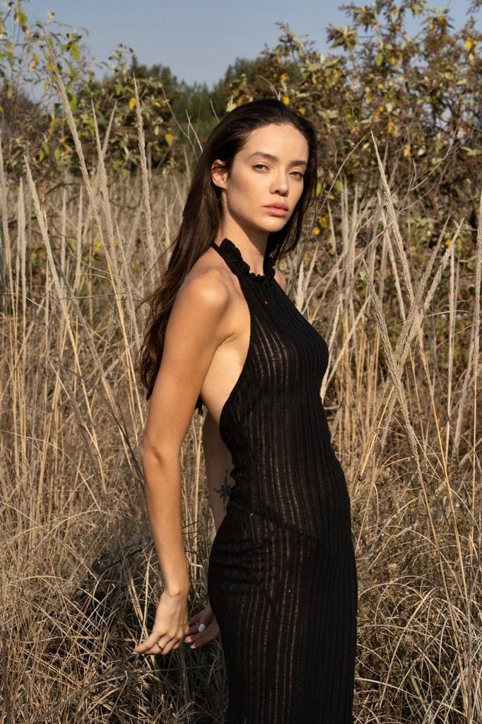 A woman in a black halter-neck dress stands in a field of tall dry grass, body angled sideways while facing the camera.