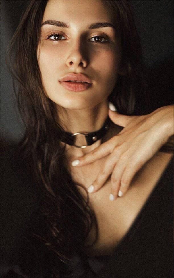 Close-up portrait against a dark background, wearing a black choker and low-cut top, hand near collarbone.