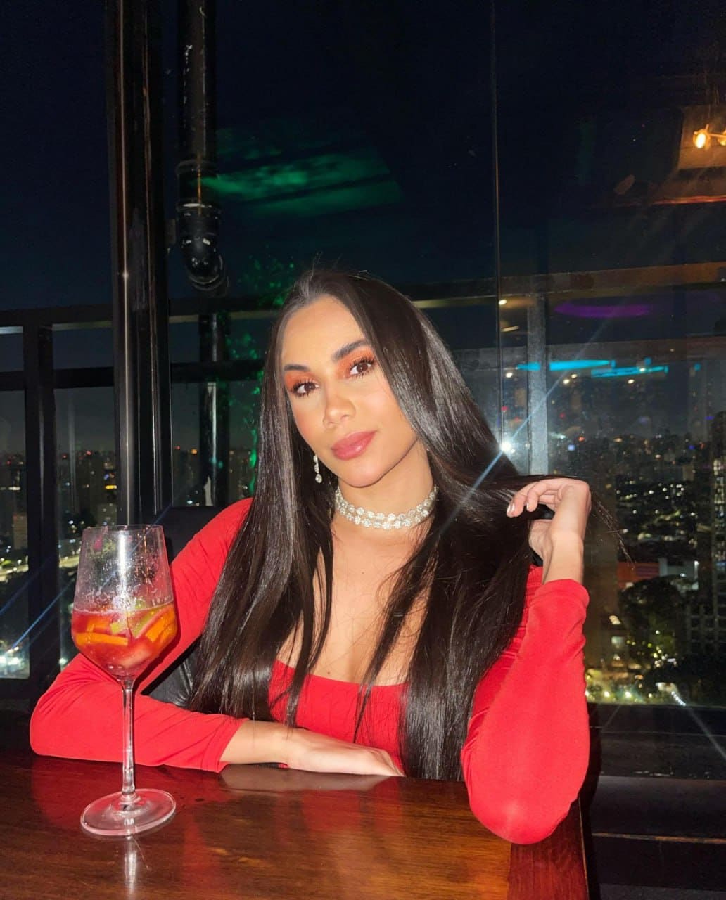 Alicia sits at a wooden table in a dim room with a night city view, wearing a red top and pearl choker, touching her hair near a cocktail.