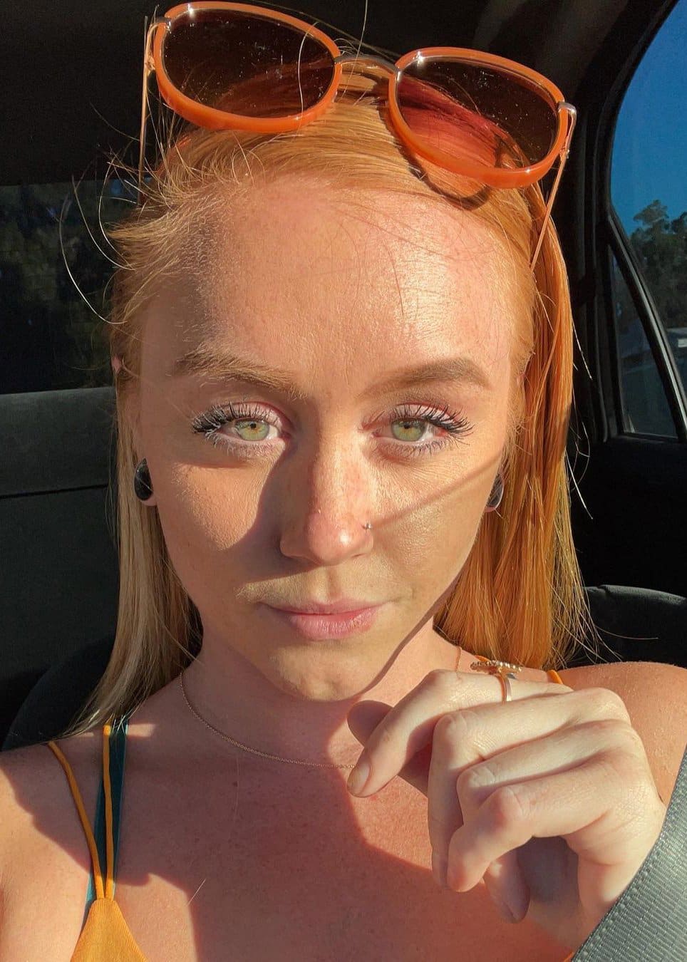 Red-haired woman with sunglasses on head sits in a car wearing a yellow and green strap top.
