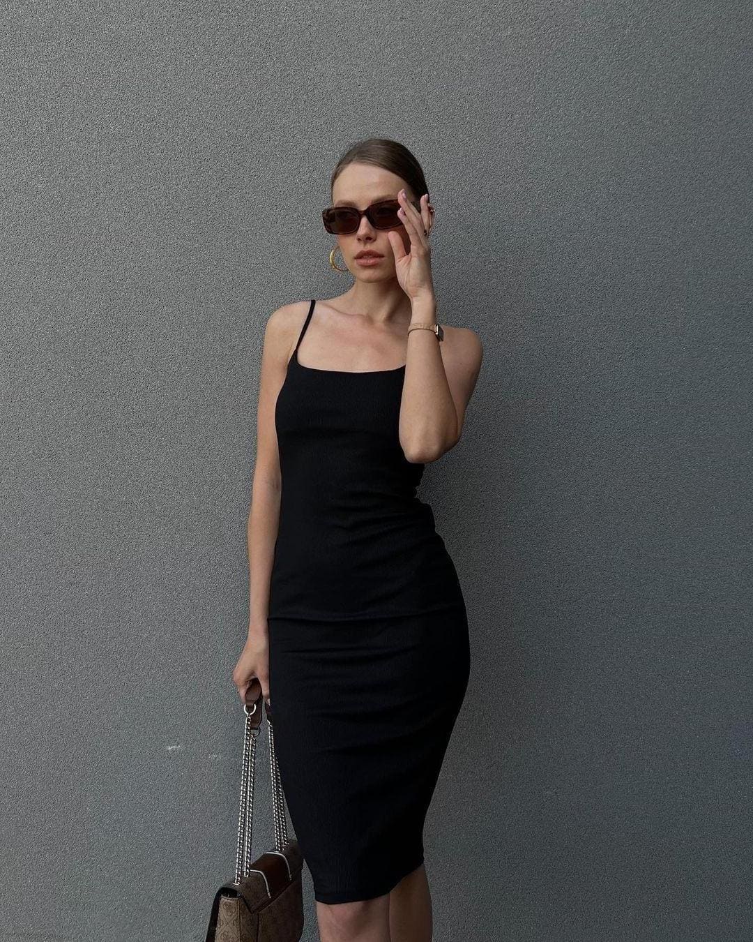 Against a gray wall, she wears a black midi dress and sunglasses, holding a chain-strap bag and touching her face.